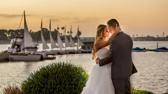 A couple in wedding attire embraces by the waterfront at sunset, with sailboats and palm-lined docks in the background.