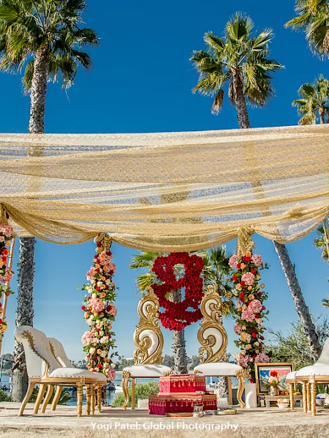 An outdoor wedding stage with floral arches, an ornate backdrop, gold chairs, and a canopy against tall palm trees.