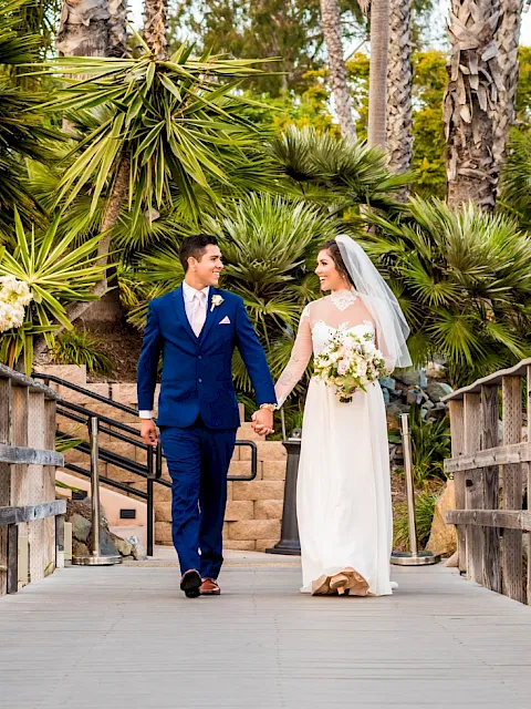 A bride and groom in blue and white walk hand-in-hand on a sunny boardwalk among palm trees and wedding flowers.