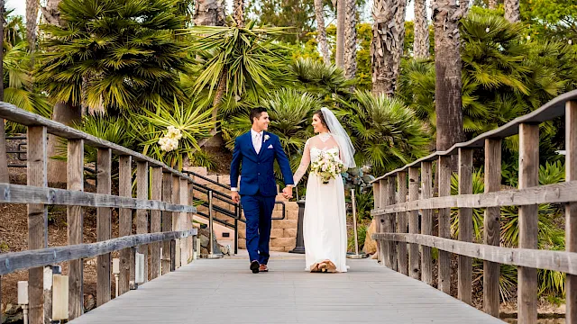A bride and groom in blue and white walk hand-in-hand on a sunny boardwalk among palm trees and wedding flowers.