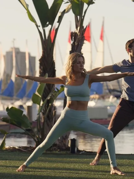 A couple practices yoga on a grassy waterfront with palm trees and sailboats in the background.
