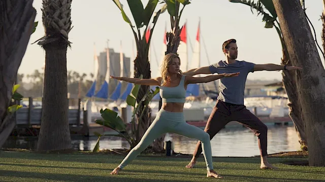 A couple practices yoga on a grassy waterfront with palm trees and sailboats in the background.