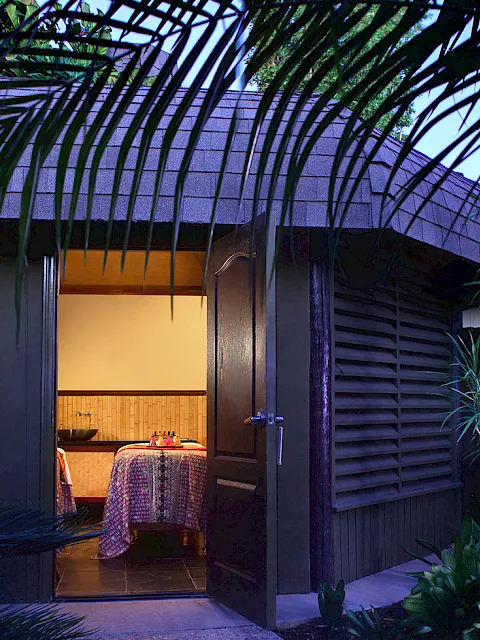 A tropical courtyard with lush plants, an arched doorway, and a small dining table set for two under warm lights.