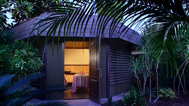 A tropical courtyard with lush plants, an arched doorway, and a small dining table set for two under warm lights.