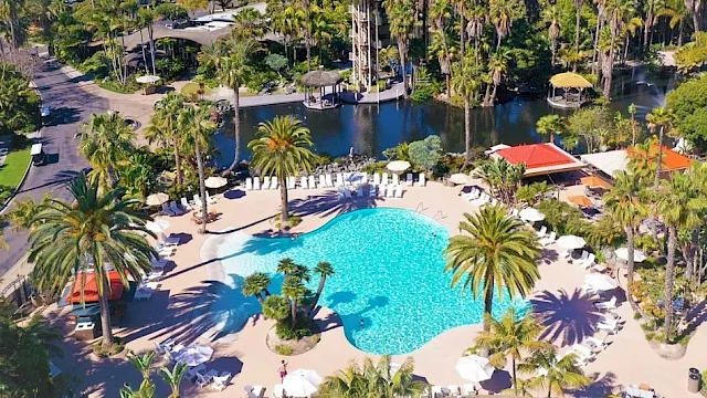 Aerial view of a tropical resort with a large swimming pool, palm trees, cabanas, and a marina by the ocean.
