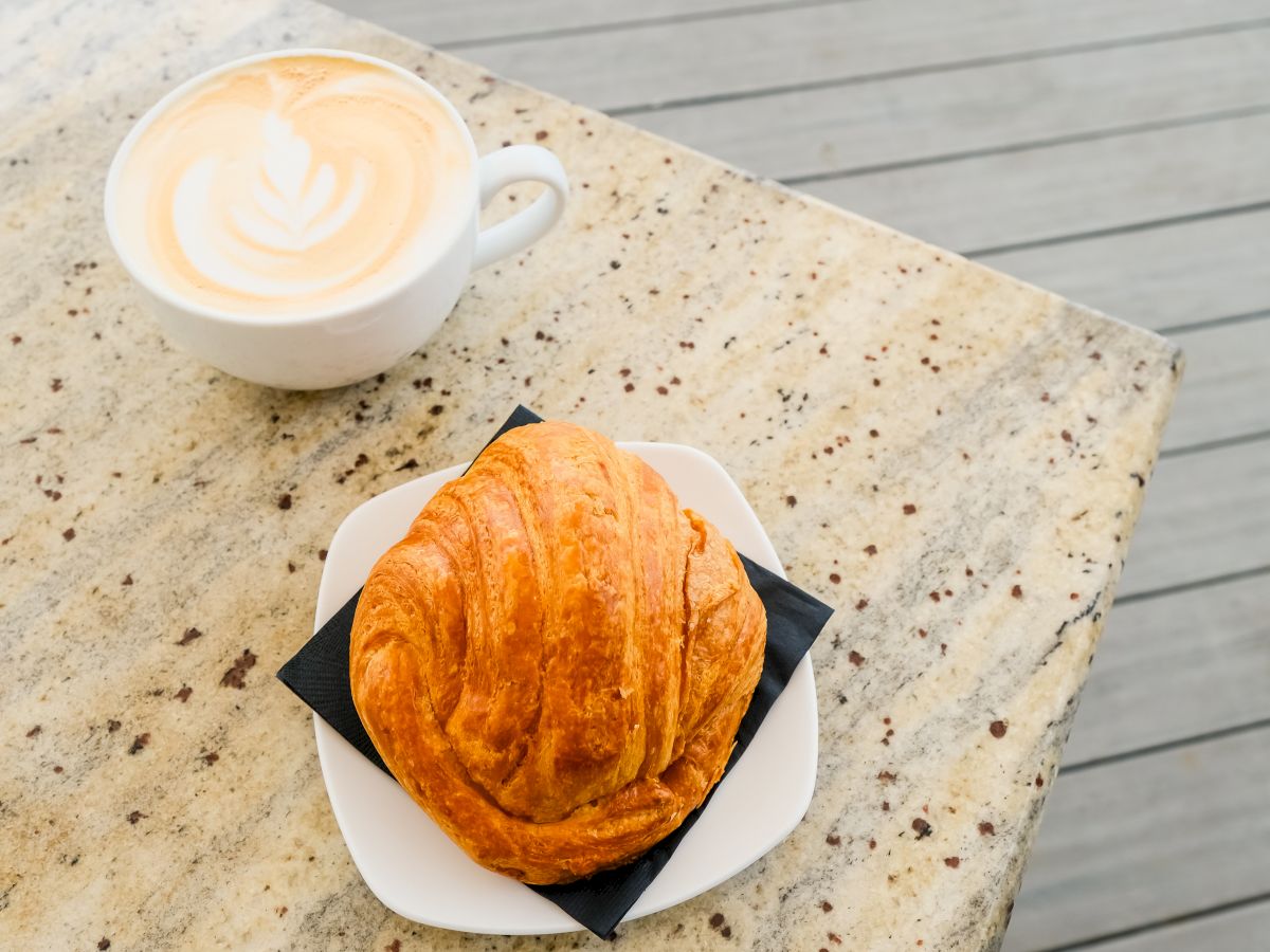 A cup of latte with latte art and a warm croissant on a plate on a speckled table.