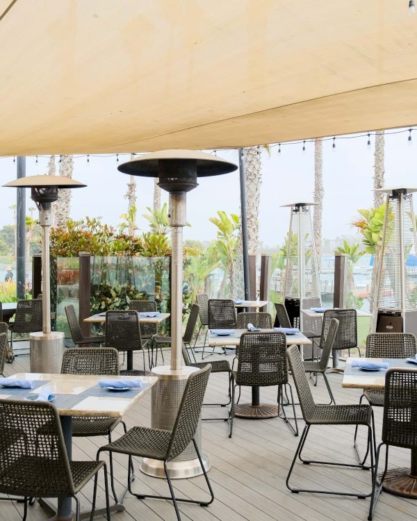 An outdoor restaurant patio with tables and chairs, tall patio heaters, and lush plants with palm trees in the background.