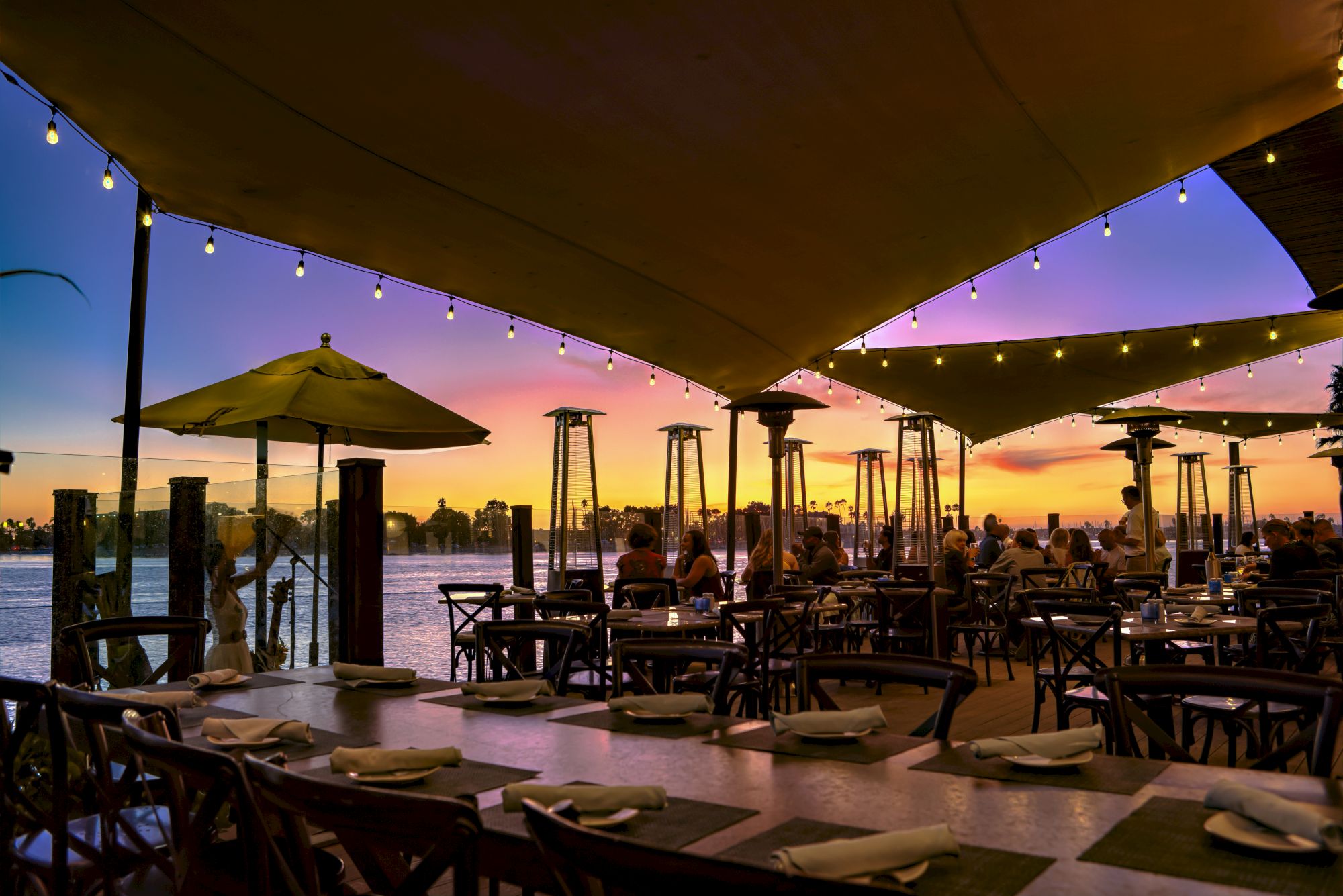 An outdoor riverside dining patio with string lights, umbrellas, and people enjoying sunset views over the water.