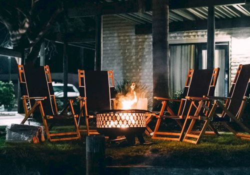 An outdoor patio with wooden lounge chairs around a flaming fire pit, with a person standing nearby in the dark.