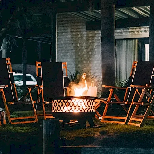 An outdoor patio with wooden lounge chairs around a flaming fire pit, with a person standing nearby in the dark.