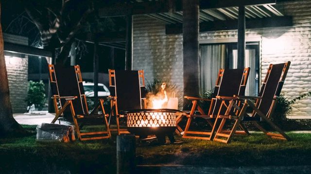 An outdoor patio with wooden lounge chairs around a flaming fire pit, with a person standing nearby in the dark.