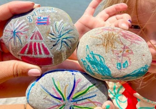 A pair of hands holds several painted rocks with colorful fireworks designs and stars along a waterfront.
