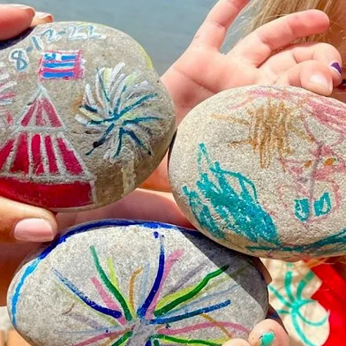 A pair of hands holds several painted rocks with colorful fireworks designs and stars along a waterfront.