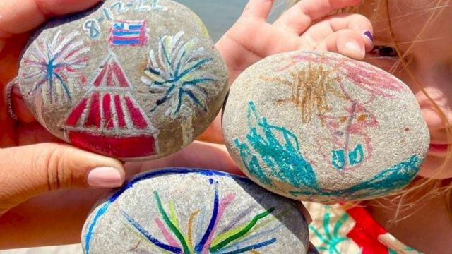 A pair of hands holds several painted rocks with colorful fireworks designs and stars along a waterfront.