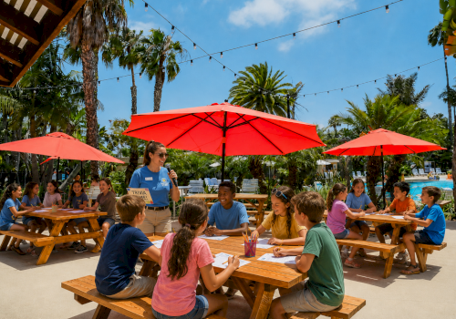 A group of kids sit at outdoor wooden tables under red umbrellas, playing a trivia game with a smiling instructor on a sunny day.