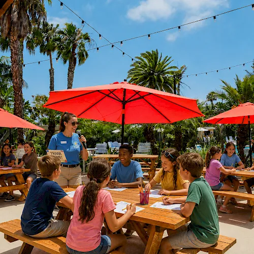 A group of kids sit at outdoor wooden tables under red umbrellas, playing a trivia game with a smiling instructor on a sunny day.