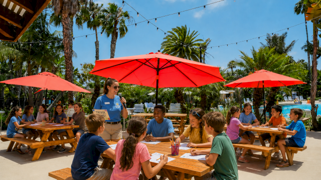 A group of kids sit at outdoor wooden tables under red umbrellas, playing a trivia game with a smiling instructor on a sunny day.
