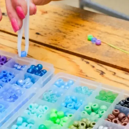 A hand is arranging colorful plastic beads in small compartment containers on a wooden table.