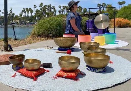 A woman in a hat stands by a seaside setup with singing bowls, colorful cushions, and a small microphone-like device.