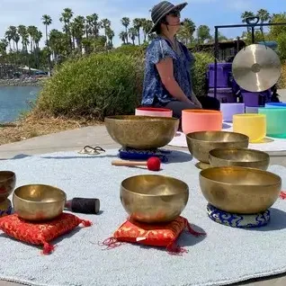 A woman in a hat stands by a seaside setup with singing bowls, colorful cushions, and a small microphone-like device.