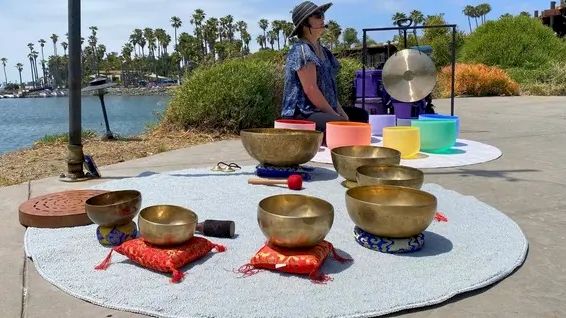 A woman in a hat stands by a seaside setup with singing bowls, colorful cushions, and a small microphone-like device.