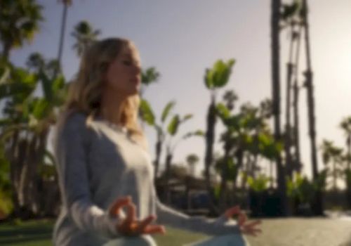 A woman in a sunny park stretches outdoors with palm trees lining the walkway behind her.