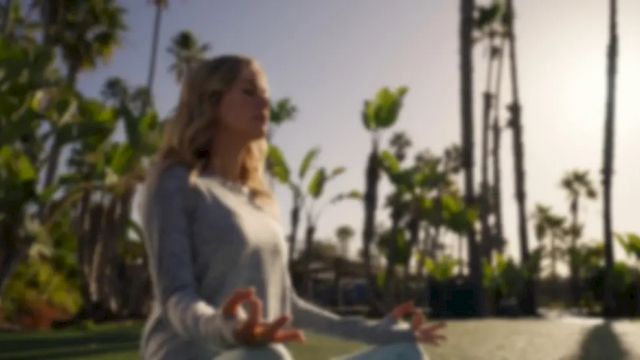 A woman in a sunny park stretches outdoors with palm trees lining the walkway behind her.