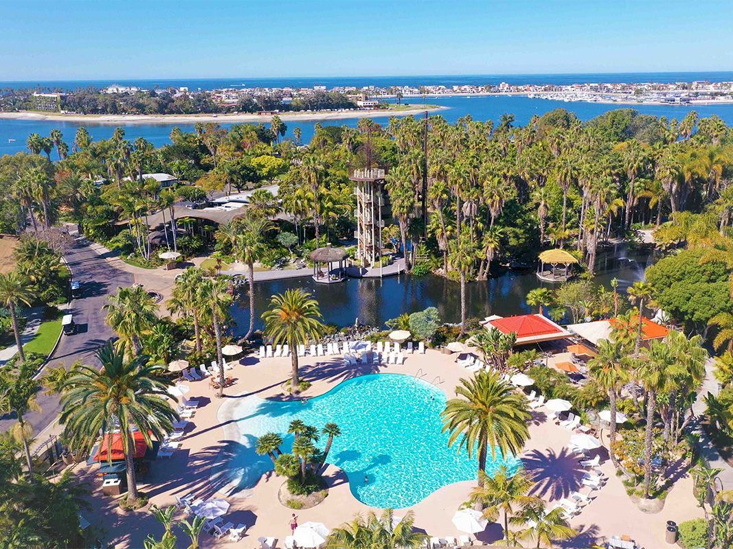 Aerial view of a tropical resort with a bright blue pool, palm trees, and canals overlooking the ocean.