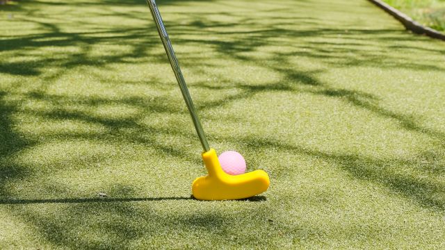 A person&rsquo;s hand holds a putter as a pink golf ball rests on short grass beside a winding putting green.