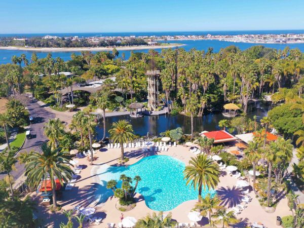 Aerial view of a tropical beach resort with a turquoise pool, palm trees, lawns, and a bay with boats and city shoreline.