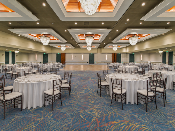 A spacious banquet hall set with round dining tables, white linens, gold-rimmed glassware, and hanging crystal chandeliers.