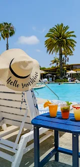 Poolside lounge by palm trees with a sun hat reading &ldquo;Hello sunshine&rdquo; and colorful drinks on a blue table.