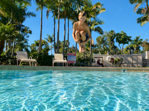 A boy leaps into a sunny pool surrounded by palm trees and poolside loungers, looking carefree.