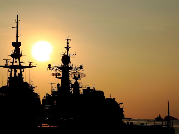 Silhouetted naval vessels at harbor with radar masts against a glowing sunset sky.
