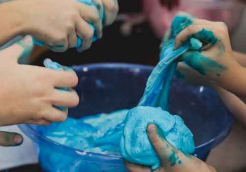 A person&rsquo;s hands are covered in bright blue slime or paint, squishing and smearing it during a messy activity.