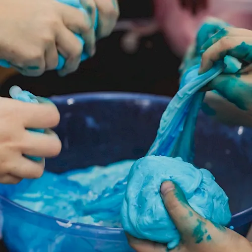 A person’s hands are covered in bright blue slime or paint, squishing and smearing it during a messy activity.