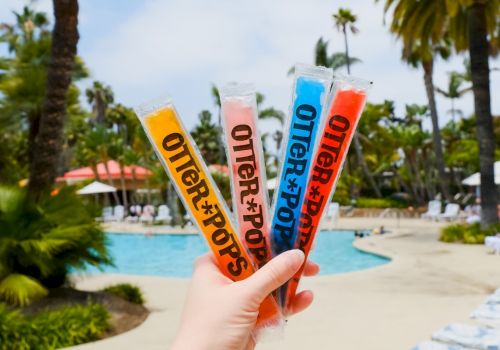 Hand holds four colorful flavored icy strips by a sunny pool with palm trees in the background.