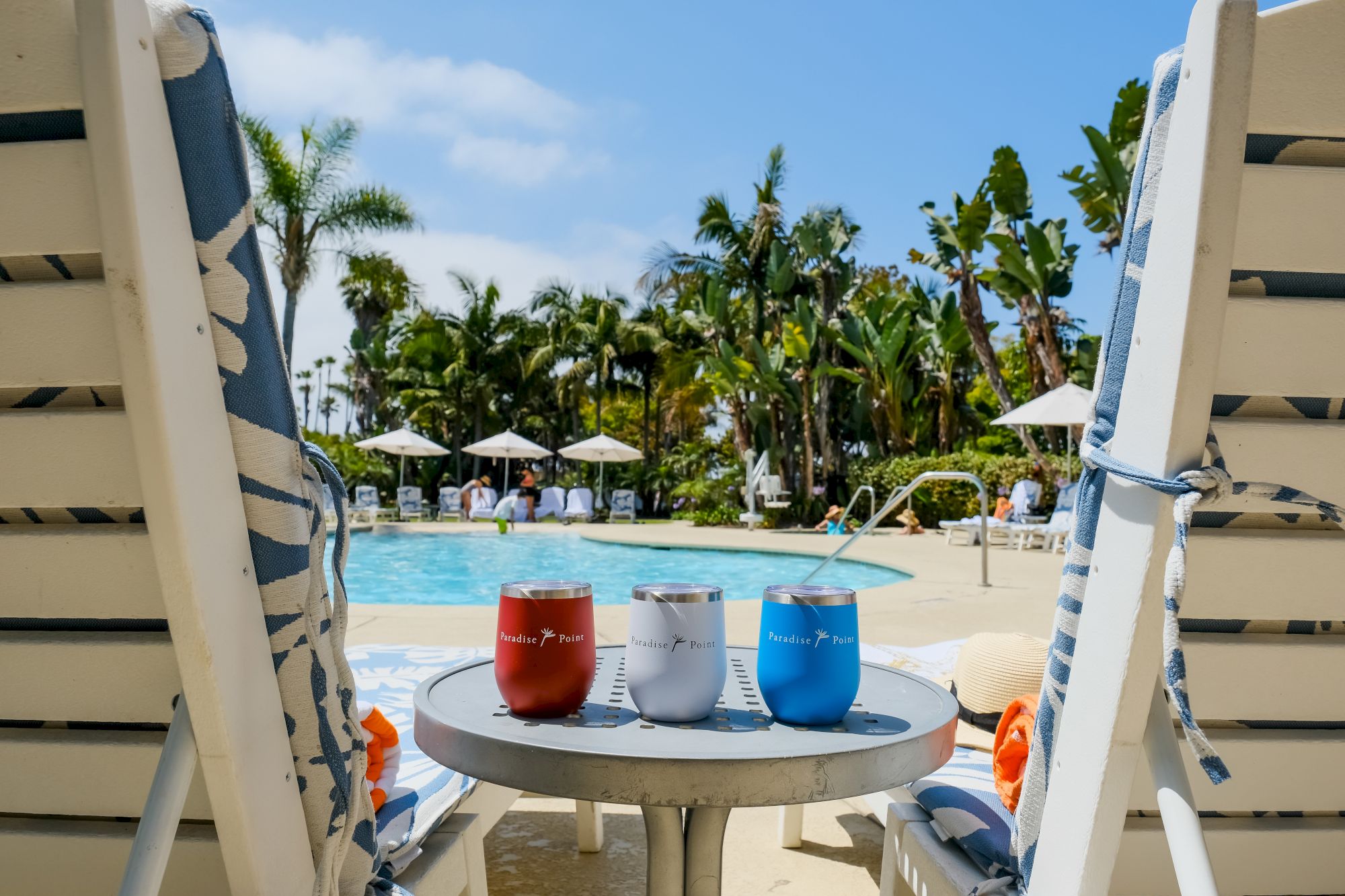 Tropical poolside view with umbrellas and palm trees, seen from a balcony with three insulated tumblers on a table.