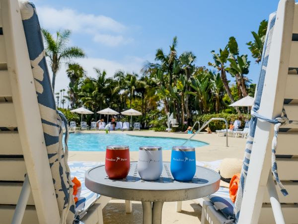 A tropical resort pool scene with palm trees, sun umbrellas, and a table of colorful drink cups by the water.