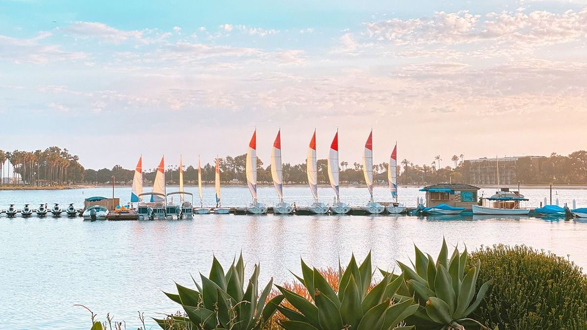 A calm lake at sunset with a line of sailboats moored on docks and palm trees along the shore.