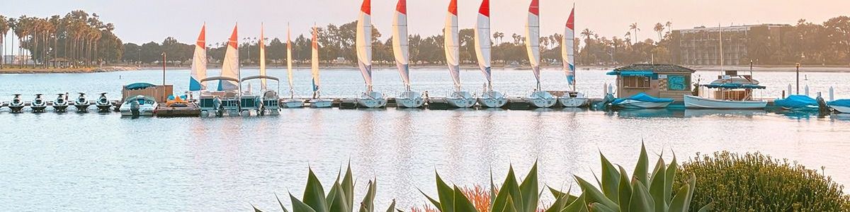 A calm lake at sunset with a line of sailboats moored on docks and palm trees along the shore.