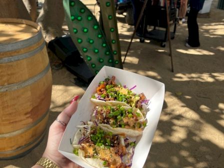 A hand holds a white tray of street tacos topped with chopped green onions and purple slaw outside in a sunny patio.