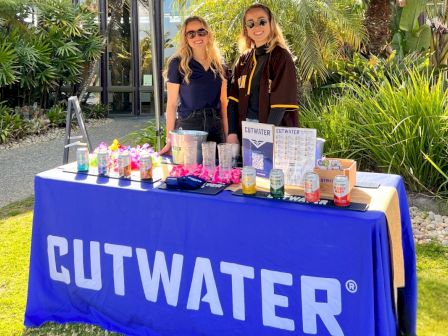 Two women stand behind a CUTWATER booth table with drinks and colorful bottles in a sunny garden.