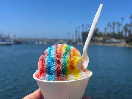 A rainbow shaved-ice snow cone with a spoon, held by the seaside under a blue sky.