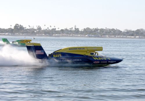 A &ldquo;Spirit of the Navy&rdquo; US Navy speedboat races across water, kicking up spray with a shoreline in the background.