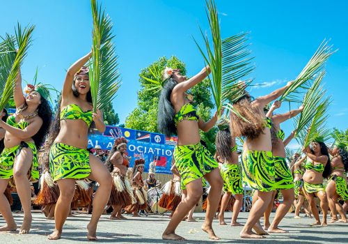 Festival dancers in green grass skirts and bikinis, lifting palm fronds, performing outdoors on a sunny day.