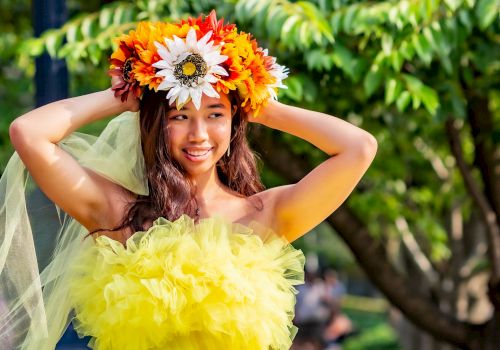 A girl in a bright green-yellow tulle dress wears a colorful flower crown outdoors, arms up and smiling.