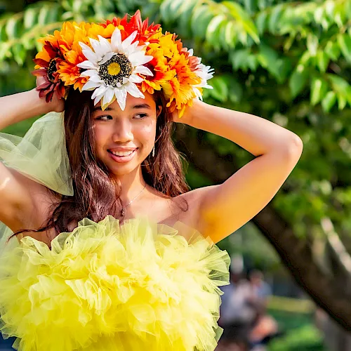 A girl in a bright green-yellow tulle dress wears a colorful flower crown outdoors, arms up and smiling.