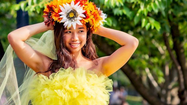 A girl in a bright green-yellow tulle dress wears a colorful flower crown outdoors, arms up and smiling.