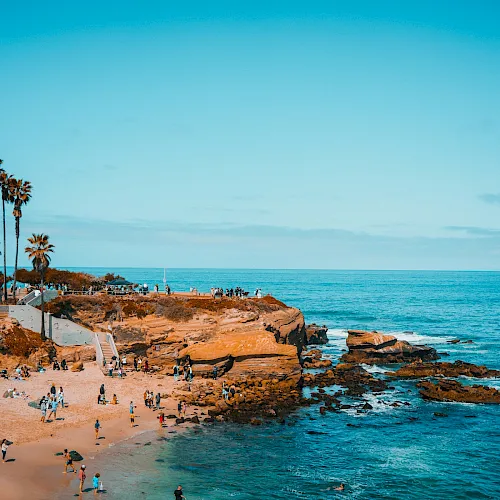 A sunny rocky coastline with palm trees, waves crashing, and people relaxing on the sandy beach.